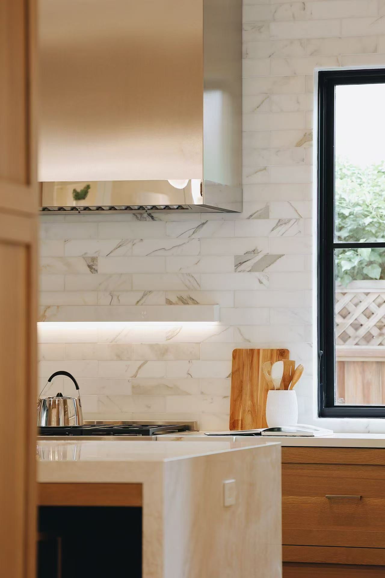 Cooking wall with stainless steel hood, stone backsplash, timber cabinet, and black-framed window