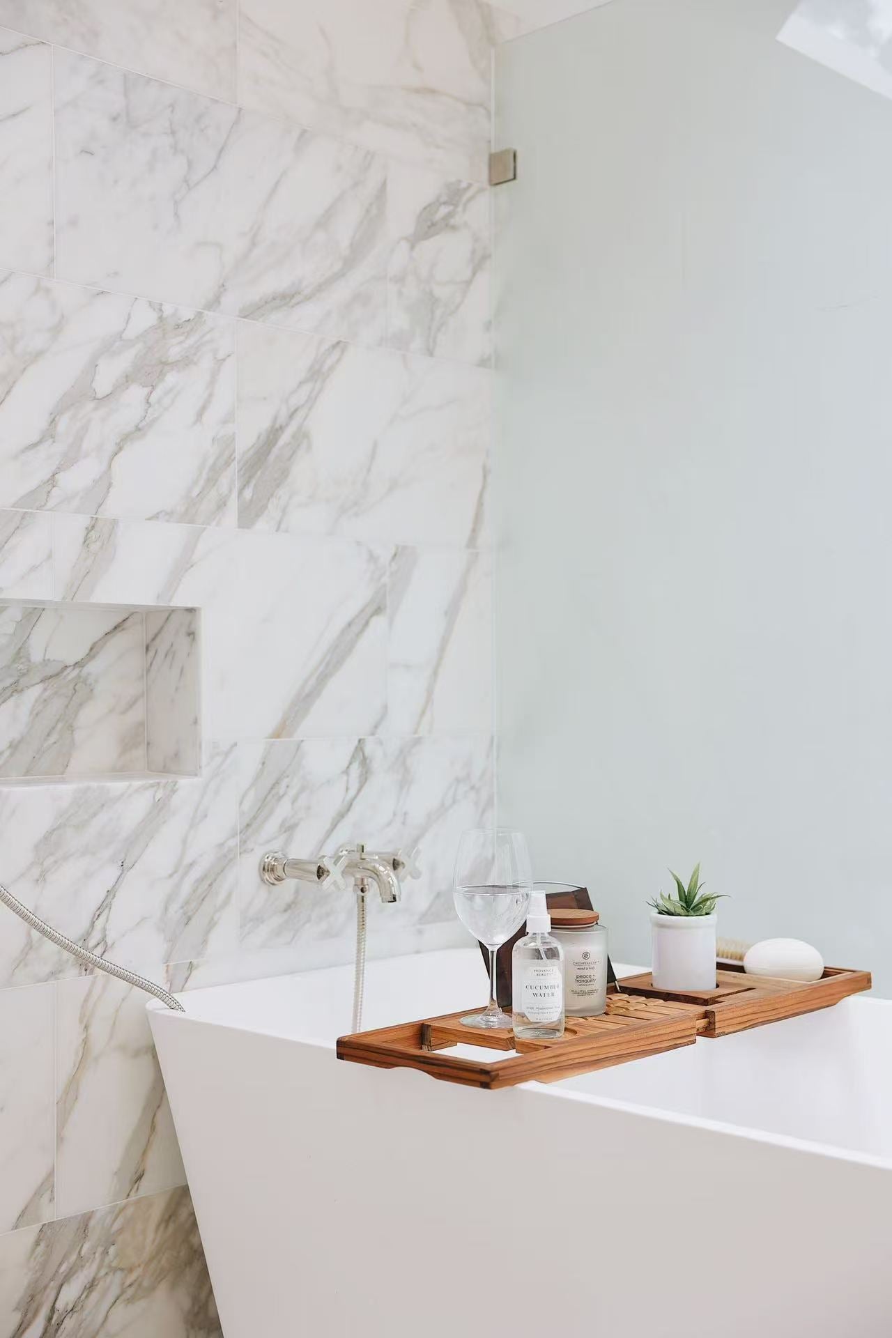 Freestanding bathtub with marble-effect wall, glass panel, and warm wood accessory