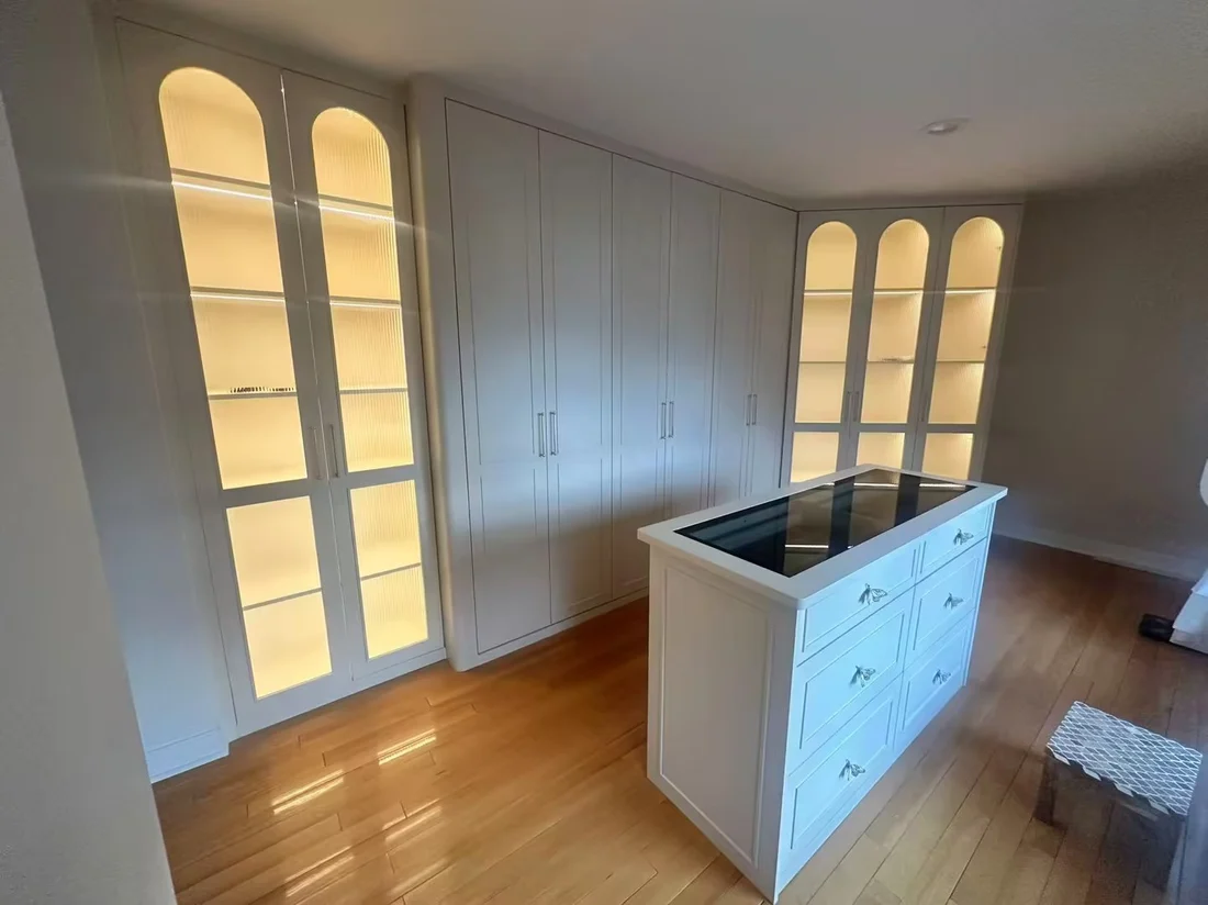 Completed New Jersey dressing room with cream lacquered cabinetry, lit arched fluted-glass display units and a stone-topped island