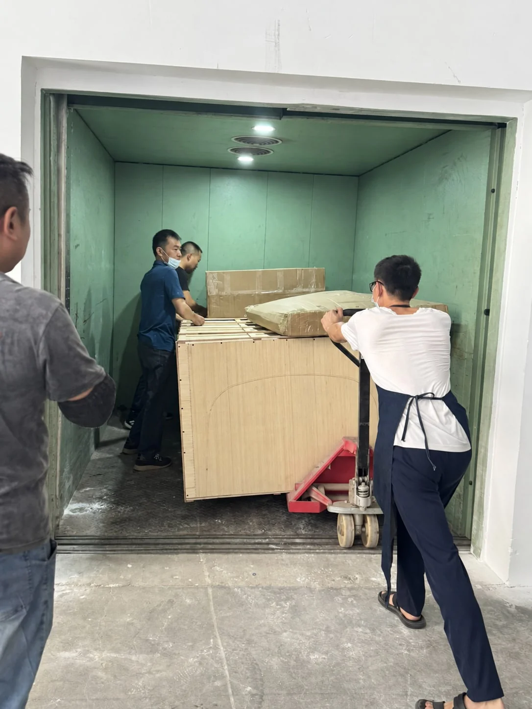 Workers moving crated furniture into a goods lift with a pallet truck